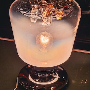 Op de afbeelding: Een vintage tafellamp met een heldere, afgeronde glazen kap en een chromen voet. De kap heeft een matte uitstraling aan de onderkant, die overgaat in helder aan de bovenkant. Een verlichte gloeilamp is zichtbaar binnenin, die een warme gloed afgeeft. De voet is rond en reflecterend.