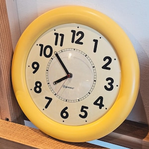 Puede incluir: Un reloj de pared redondo con un marco amarillo brillante y una esfera de color crema. El reloj presenta números arábigos negros en negrita del 1 al 12, manecillas negras para las horas y los minutos, y el logo de Guzzini en el centro.