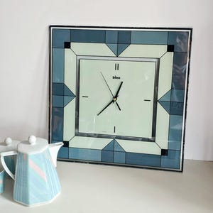 Può includere: Un orologio da parete quadrato con quadrante verde pallido, lancette nere e il marchio bino stampato in nero. La cornice presenta un bordo geometrico di pannelli di vetro blu e verde chiaro con accenti neri. Accanto si trova una teiera in ceramica bianca.
