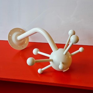 Può includere: Un attaccapanni bianco in stile moderno della metà del secolo. Ha una piastra di montaggio circolare e un braccio curvo. L'attaccapanni presenta una sfera centrale con più pomelli sporgenti, creando un design a forma di stella. Su sfondo rosso e bianco.