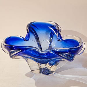 Könnte beinhalten: Eine dekorative Glasschale mit leuchtend blauem Innenraum. Die Schale hat eine einzigartige, organische Form mit fließenden Kurven und abgerundeten Kanten. Die Farbe geht von tiefblau in klar über und erzeugt einen optisch ansprechenden Kontrast. Die Schale ist wahrscheinlich für dekorative Zwecke.