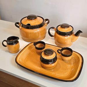 Puede incluir: Un juego de té de cerámica con un esmalte brillante color miel y bordes de color marrón oscuro. El conjunto incluye una tetera con tapa, un azucarero con tapa, una sopera con tapa, una jarra de leche, dos tazas pequeñas y una bandeja rectangular.