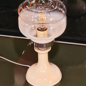 Può includere: Una moderna lampada da tavolo con paralume in vetro trasparente e una lampadina a luce calda. La lampada ha una base color crema e un accento metallico cromato. Il design è elegante e contemporaneo, adatta per un soggiorno o una camera da letto.