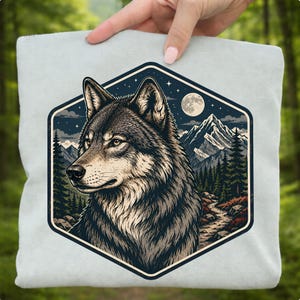 May include: A light blue sweatshirt featuring a detailed illustration of a wolf within a hexagonal frame. The artwork depicts a wolf's head, mountains, a full moon, and a forest scene, creating a nature-themed design.