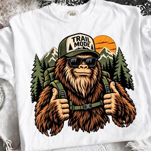 May include: A white t-shirt featuring a graphic of a brown, furry creature wearing sunglasses and a green cap with the text "TRAIL MODE" printed on it. The creature has a backpack, gives a thumbs up, and stands before mountains and trees.