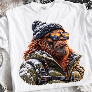 May include: A white t-shirt featuring a graphic of a brown, furry creature wearing a dark blue knit beanie, orange-tinted sunglasses reflecting a mountain sunset, and an olive green quilted winter jacket covered in white snow and black straps.