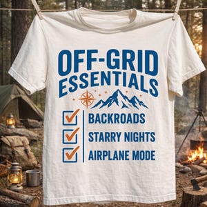 Może przedstawiać: Kremowa koszulka z niebieskim napisem "OFF-GRID ESSENTIALS", kompasem, górami i polami wyboru dla "BACKROADS", "STARRY NIGHTS" i "AIRPLANE MODE". Koszulka wisi na sznurku do prania.