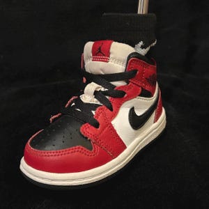 May include: A red, black, and white baby trainer with black laces and a black Nike swoosh. The high-top shoe has a white sole and a red collar. A black sock is inside the shoe.
