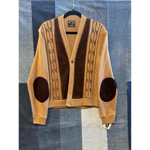 Pode incluir: Um cardigan bege com detalhes em camurça marrom e cotoveleiras. O suéter apresenta um design de malha de cabo nos painéis frontais e mangas, com fecho de botão. Está pendurado em um cabide preto.