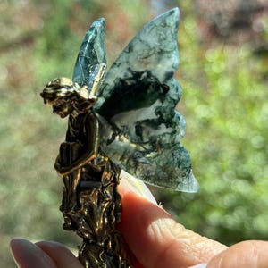 May include: A small figurine of a seated figure with a metallic bronze-toned body and translucent moss agate wings featuring dark green, fern-like inclusions. The figure is depicted in a resting pose with detailed textures on the base and wings.