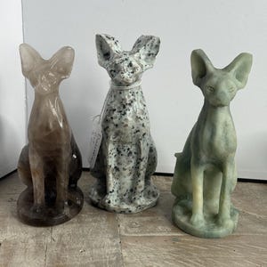 May include: Three stone cat sculptures in a row. The cats are sitting upright with large ears. The cat on the left is a translucent brown color. The middle cat is speckled gray and white. The cat on the right is a pale green color.