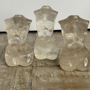 May include: Three translucent crystal torso sculptures. The sculptures are carved in the shape of a female torso. The crystals have a light, neutral color and sit on a weathered wooden surface. The sculptures are displayed in a row.