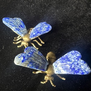 May include: Two insect-shaped decorative ornaments featuring metallic bronze-toned bodies and wings crafted from polished blue lapis lazuli stone with natural white and gold-colored flecks, displayed in a top-down view with visible segmented legs.