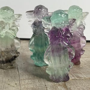 May include: A collection of carved angel figurines in various colors, including green, purple, and clear. The angels have detailed wings and are posed with hands clasped in prayer. The figurines are made of a translucent material.