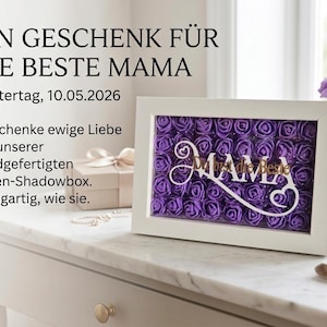 May include: A white framed shadowbox filled with purple roses. The word "MAMA" is displayed in a decorative font. The text "EIN GESCHENK FÜR DIE BESTE MAMA" is at the top, with additional text below.