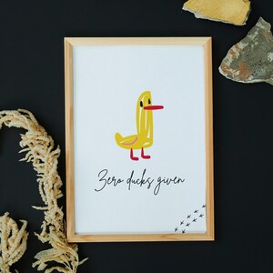 May include: A framed print featuring a whimsical yellow duck illustration with a red beak and feet. The phrase "Zero ducks given" is written below the duck in a cursive font. The artwork is set against a white background.