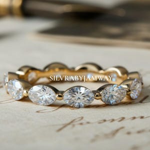 Op de afbeelding: Een gouden ring met een reeks ovale, heldere edelstenen. De ring ligt op een vintage papieren achtergrond. De tekst "SILVRABYJAMWAY" is zichtbaar. Een sieraad.