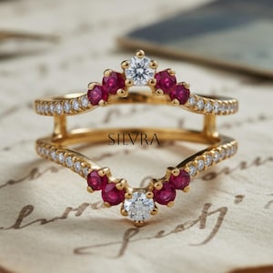 Puede incluir: Un anillo dorado con un diseño en forma de V, con pequeñas piedras rojas y diamantes transparentes. El anillo tiene la palabra "SILVRA" grabada. El anillo está sobre un fondo de papel vintage.