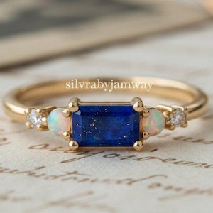 May include: A gold ring featuring a rectangular, deep blue gemstone, flanked by two smaller opalescent stones and tiny diamonds. The ring is set on a vintage paper background. The text "silvrabyjamway" is visible on the ring.