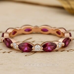 May include: Rose gold ring with alternating marquise-cut red gemstones and small round white gemstones. The ring is set on a vintage-style paper background with a gold pen in the background. The ring is a continuous band.