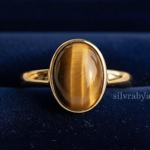 May include: A gold ring with an oval tiger's eye stone. The ring has a polished gold band and a bezel setting that holds the brown and gold stone. The ring is displayed in a dark blue velvet ring box.