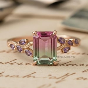 May include: A rose gold ring with a rectangular, bi-colour pink and green gemstone. The band features small, purple gemstones and a leaf design. The ring is set on a vintage paper background.