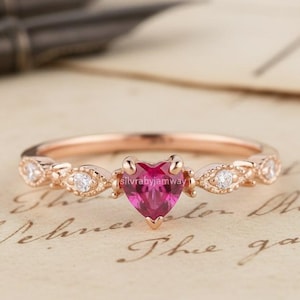 May include: A rose gold ring with a heart-shaped, magenta-colored gemstone at the center. The band features small, clear gemstones and decorative accents. The ring is set against a vintage paper background.