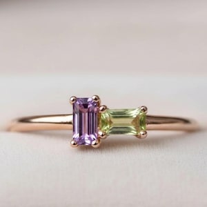 Op de afbeelding: Een roségouden ring met twee rechthoekige edelstenen: een paarse en een groene. De edelstenen zijn naast elkaar geplaatst. De ring wordt gepresenteerd in een witte doos. Het ontwerp is elegant en modern. Een prachtig sieraad.