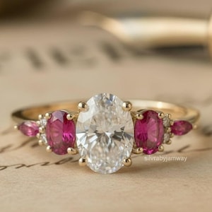 Può includere: Un anello d'oro con una grande pietra centrale di diamante taglio ovale, affiancata da due pietre preziose di rubino taglio ovale. Piccoli accenti di rubino e diamante su entrambi i lati dei rubini. L'anello è posto su uno sfondo di carta vintage.