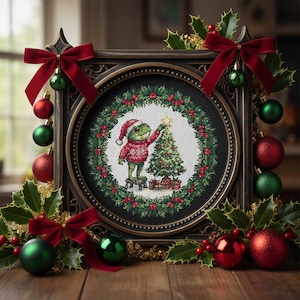 Könnte beinhalten: Ein gerahmtes Kreuzstichbild mit einem Frosch, der einen Weihnachtsbaum schmückt, umgeben von einem Kranz aus Stechpalmen und roten Beeren. Der Rahmen ist mit roten Schleifen und roten und grünen Kugeln verziert. Das Bild befindet sich auf einer Holzoberfläche.