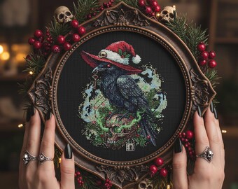 May include: A framed cross-stitch featuring a black raven wearing a Santa hat, surrounded by a decorative frame with skulls, red berries, and greenery. The raven is perched on a skull, with a dark background.