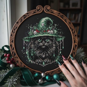 May include: A cross-stitch of a black cat wearing a green witch hat adorned with skulls and red berries, set in a round, ornate wooden frame. The cat has green eyes and is surrounded by green foliage. The frame is decorated with swirling patterns.