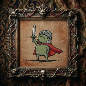 Puede incluir: Obra de arte de punto de cruz de un caballero rana verde sosteniendo una espada, con un casco y una capa roja. La obra está enmarcada en un marco de madera ornamentado con detalles de calaveras y espinas. El fondo es de tela texturizada.