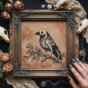 Puede incluir: Obra de arte de punto de cruz de un cuervo con diseño de esqueleto, posado en una rama con flores rojas. El cuervo está enmarcado en un marco de madera oscura, con un fondo beige con una luna y estrellas.