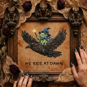 Może przedstawiać: Haft krzyżykowy w ciemnej drewnianej ramie przedstawiający zielonego żabiego czarodzieja jadącego na czarnym kruku, trzymającego różdżkę. Tekst "WE RIDE AT DAWN" znajduje się pod obrazem. Ozdobne posągi gargulców u góry ramy.