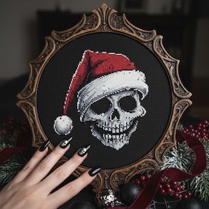 May include: Cross-stitch artwork of a skull wearing a Santa hat, displayed in a decorative wooden frame. The skull is white with a detailed design, set against a black background. The Santa hat is red and white. The frame is brown.