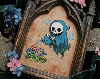 May include: Cross-stitch artwork in a dark wooden frame. The image shows a cartoon grim reaper watering flowers. The reaper is wearing a blue cloak and has a skull face. The flowers are pink, blue, and yellow.