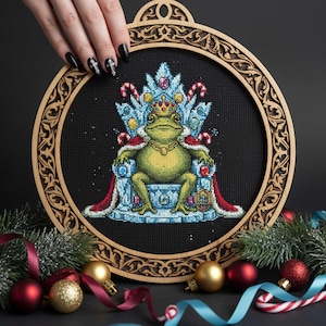 May include: Cross-stitch embroidery of a regal frog wearing a crown and sitting on a throne. The frog is green and gold, with a red and blue throne. The embroidery is set in a round wooden frame with decorative edges. Christmas ornaments and greenery are in the background.