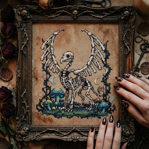 May include: Cross-stitch artwork in a dark, ornate frame. The piece depicts a skeletal griffin with outstretched wings, surrounded by stylized mushrooms, vines, and crescent moons. The color palette includes beige, black, blue, and green.