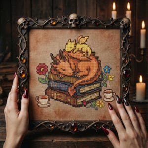 May include: Cross-stitch art of a sleeping orange dragon and a smaller yellow dragon on a stack of books. The artwork is framed in a dark, ornate frame with skulls and jewels. Flowers and teacups are also part of the design.
