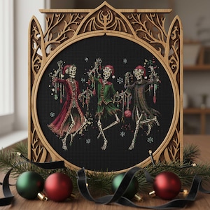 May include: A framed cross-stitch featuring three dancing skeletons in festive attire, each wearing a Santa hat and holding ornaments. The artwork is set against a black background and framed in a decorative wooden frame. Christmas ornaments and greenery are arranged at the base.