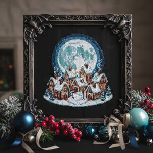 Könnte beinhalten: Ein gerahmtes Kreuzstichbild, das eine Winterdorf-Szene unter einem Vollmond darstellt. Das Kunstwerk zeigt schneebedeckte Dächer, einen großen blauen Mond und ist in einem dekorativen, dunkel getönten Rahmen mit Fledermausflügel-Ecken eingefasst. Weihnachtsornamente umgeben den Rahmen.