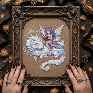May include: A framed cross-stitch artwork depicts a fairy riding a cloud-like dragon, reading a book. The fairy has purple hair and wings, wearing a pink dress. The artwork is in a decorative, ornate brown frame, with a neutral background.