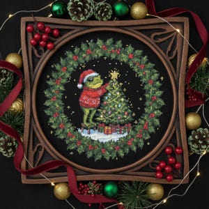 May include: Cross-stitch art of a frog decorating a Christmas tree, wearing a Santa hat and sweater. The artwork is framed in a dark wood square frame, surrounded by a wreath of holly and berries. Christmas ornaments and ribbon complete the festive scene.