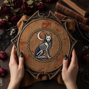 This gothic cross-stitch pattern features a skeletal fox seated beneath a glowing crescent moon, surrounded by a decorative Celtic-style ring and shimmering stars. The fox’s elegant bone structure is rendered in soft ivory and pale gray tones, contrasted against a deep blue night sky dotted with golden starlight. The circular framing gives the design a symbolic, talisman-like presence, making it perfect for gothic décor, fantasy art collections, and celestial-themed embroidery displays.