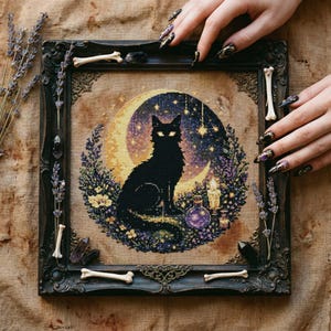 Op de afbeelding: Een ingelijste kruissteekafbeelding met een zwarte kat op een maan, omringd door sterren, bloemen en een flesje. Het donkere frame is versierd met botvormige accenten en kristallen. Een mystiek kunstwerk.