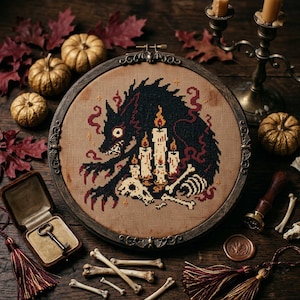 May include: A circular embroidery hoop frame containing a cross-stitch design of a black wolf surrounded by lit candles and bones on tan fabric. The dark wooden frame features ornate metal accents with small bat figures at the top and bottom.