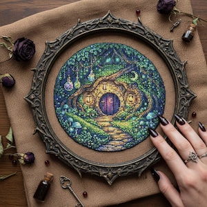 May include: A detailed cross-stitch artwork of a whimsical hobbit home, framed in an ornate, dark metal frame. The scene features a round door, lush greenery, and glowing lanterns. The artwork is set on a burlap-like background.