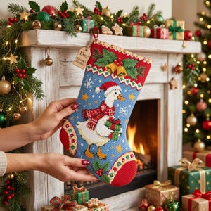Op de afbeelding: Een kerstkous met een kruissteekontwerp van een witte gans met een rode muts en sjaal die een groen cadeau vasthoudt. De blauwe achtergrond bevat sneeuwvlokken en sterren, met een houten label met de tekst Etsy Auntie's Stitches.