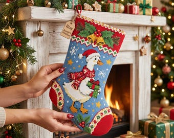 May include: A Christmas stocking featuring a cross-stitched design of a white goose wearing a red hat and scarf while holding a green gift. The blue background includes snowflakes and stars, with a wooden tag labelled Etsy Auntie's Stitches.